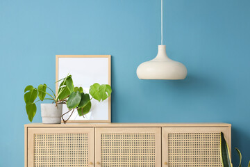 Chest of drawers with houseplant, lamp and frame near blue wall in room. Closeup