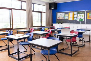 Empty classroom with desks and chairs, laptop on one desk, bright windows, copy space
