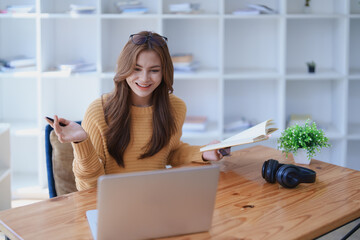 Smiling young woman engaged in a joyful online e-learning video call, gestu while holding a book...