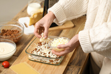 Beautiful mature woman adding cheese to delicious lasagna at table in kitchen, closeup