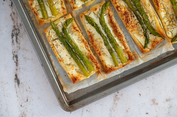 Small puff pastry tarts with green asparagus, fresh cream and parmesan. Asparagus pie with cheese on a metal tray on stone background. Spring healthy food.