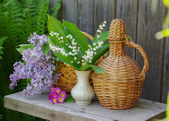 A bouquet of lilies of the valley, a bunch of lilacs, a bottle of wine on a wooden garden bench.
