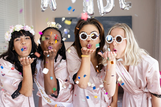 Diverse female friends celebrating bachelorette party, wearing robes and blowing confetti, at home