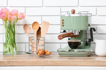 Modern coffee machine with glass cup of hot espresso and plate of sweet cakes on counter near white brick wall in kitchen