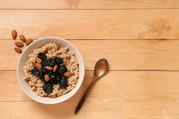 Bowl of tasty oatmeal with prunes, almonds and spoon on light wooden background