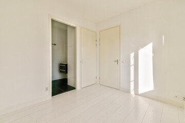A bright and spacious interior featuring white walls, light wooden flooring, and natural light casting shadows. An open doorway leads to another room.