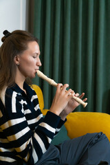 Young woman musician playing recorder flute at home, concentrating on the music with green curtain...