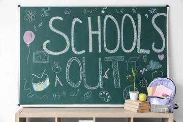 Shelf with backpack, books and text SCHOOL'S OUT on blackboard near light wall