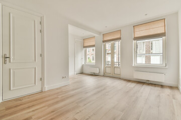 A spacious, modern living room with light-colored walls, wooden flooring, and large windows dressed with minimalistic blinds.