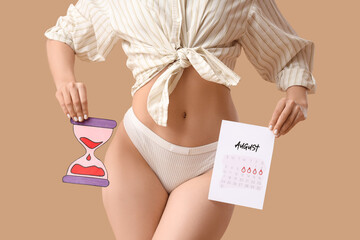 Young woman with menstrual calendar and paper hourglass on beige background