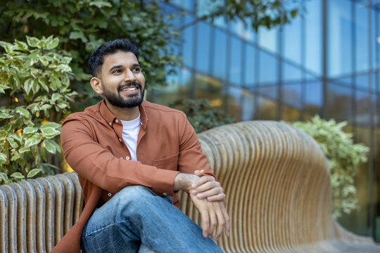 A smiling man in a casual outfit sits on a wooden bench, enjoying a sunny day outdoors with a modern building backdrop. - Powered by Adobe