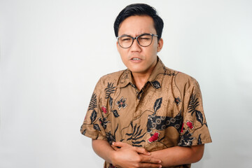 expressive asian man wearing batik and glasses with uncomfortable expression, hand on stomach due...