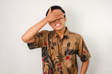 expressive asian man with sad expression, wearing batik, hands covering eyes