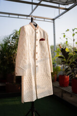 ivory sherwani with intricate embroidery, displayed on a hanger in natural daylight, perfect for Indian weddings and traditional ceremonies.