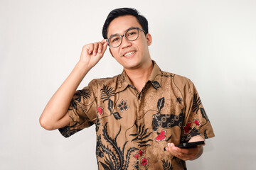 an asian man with a confident expression wearing a batik shirt and glasses holding a smartphone