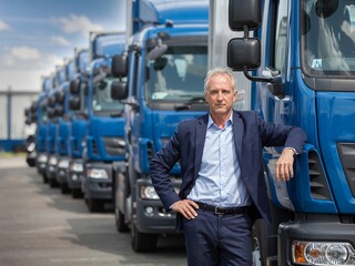 A professional portrait photograph of a confident warehouse manager standing near a fleet of blue trucks
