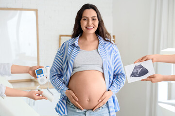 Young pregnant woman and hands holding sonogram image with fetal doppler in clinic
