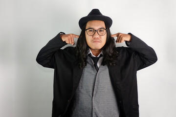 long haired asian man dressed as mafia wearing glasses and hat, making ear covering gesture with...