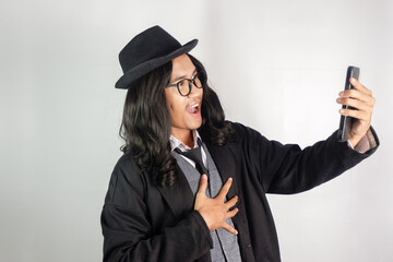 Asian man with long hair dressed as a mafia wearing glasses and a hat looking at a smartphone with...