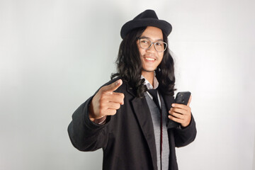 Asian man with long hair dressed as a mafia wearing glasses and a hat, holding a smartphone with a...