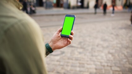 Caucasian man standing still in middle of city. View from behind shoulder of male holding smartphone with green screen. Chroma key on display. Steadily looking at mobile device. People in background.