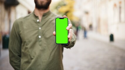 Caucasian man standing in blurred background. Showing phone to camera with one hand. Standing in middle of street. Smartphone with green screen. Chroma key on display. Advertisement.