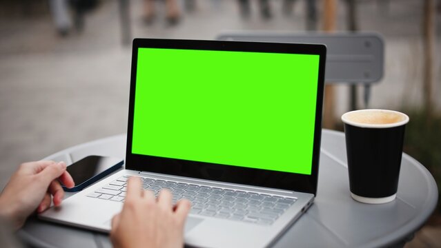 Camera focus on small table with smartphone, laptop and coffee cup. Someone using laptop with green screen. Using touchpad with one hand. Chroma key visible on display. People in blurred background.