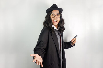Asian man with long hair dressed as a mafia wearing glasses and a hat, holding a smartphone and his...
