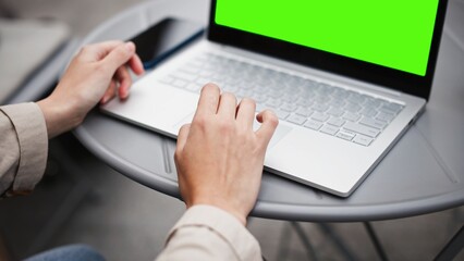 Camera closing in on Caucasian girls hand swiping on touchpad of big laptop. Using computer outside with green screen. Chroma key visible on display. Cold cloudy weather. Professional PC user.