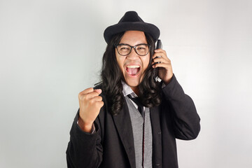 Asian man with long hair dressed as a mafia wearing glasses and a hat, making a phone call and making gestures celebrating victory