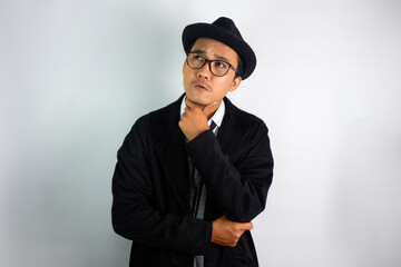 Asian man dressed as a mafia wearing glasses and a hat, making a hand gesture on his chin with a...
