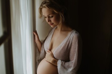 Naklejka premium A pregnant woman is looking out the window. She is wearing a white gown and has a baby bump