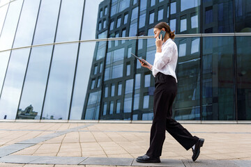 Business Professional Walks Confidently While Talking on the Phone and Using a Tablet Near Modern...
