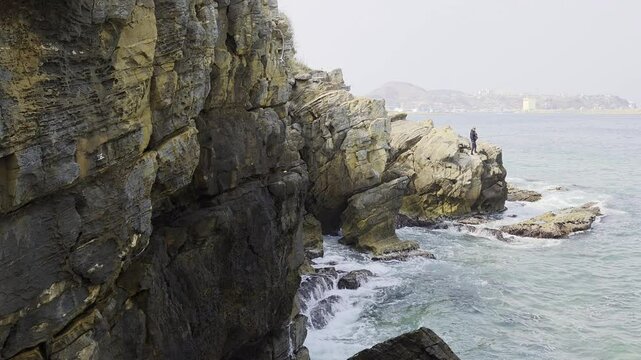A person is standing on a rocky cliff by the sea, facing the water. The cliff is made of layered rock formations, and a coastal town is visible in the distance.

