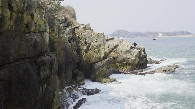 A person is standing on a rocky cliff by the sea, facing the water. The cliff is made of layered rock formations, and a coastal town is visible in the distance.