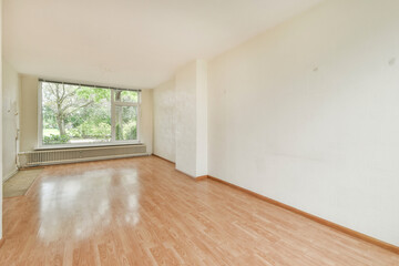 A spacious and empty room featuring a large window that opens to a natural view. The wooden floor...