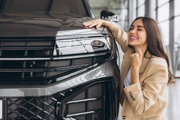 Charming young Caucasian woman hugging hood of new car and show fist and say yes in modern bright...