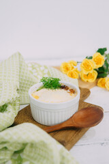 Baked casserole served in a white ramekin garnished with fresh green dill on a wooden board, accompanied by yellow roses and soft light.