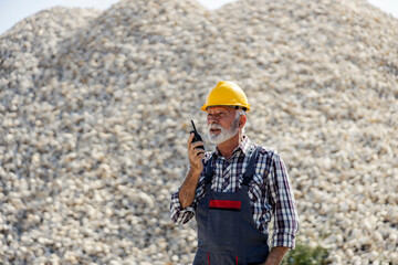 Portrait of old construction worker standing at site and talking on walkie talkie.