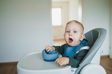 A one and a half year old boy sits on a high chair and independently eats soup from a silicone plate with a spoon. Silicone utensils for feeding children. The child learns to eat independently.