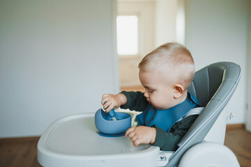 Smiling boy eats soup sitting on a chair. Hungry boy puts food in his mouth. Baby one year and three months old eats independently. Silicone utensils for feeding children.