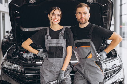 Two smiling workers, mechanic man and woman in grey uniform standing at open hood, garage vehicle shop, auto mechanic technician maintenance customer car automobile at repair service.