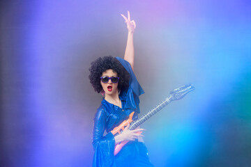 Fototapeta premium Girl comic pop star posing in black wig with toy guitar on blue background. Rock style of seventies