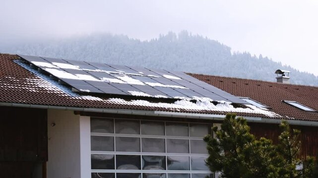 Snow melting and slipping down on modern solar panels. Snow covered roof on foggy winter day in the Bavarian Alp region. Concept of low effectivity of producing renewable electricity or clean energy.