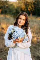 Portrait of a cute girl in a white dress barefoot in a garden with blue hydrangea. Summer walks in nature under sunlight