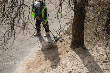 A gardener in work attire uses fan rakes to neatly clean the lawn next to a walking path in the...