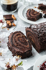 Delicious chocolate brownie roll cake stuffed with cream, cherries and nuts on light background with flowers and cup of coffee. Sweets, dessert and pastry, top view