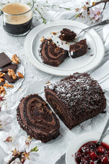 Delicious chocolate brownie roll cake stuffed with cream, cherries and nuts on light background with flowers and cup of coffee. Sweets, dessert and pastry, top view