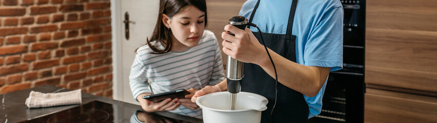 Banner of teenage brother mixing ingredients for a cake using hand blender while curious sister looking inside of the bowl and holding tablet in kitchen. Siblings learning culinary skills concept.