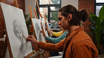 Group of adult people sitting at easels drawing pictures with pencils during master class in studio.